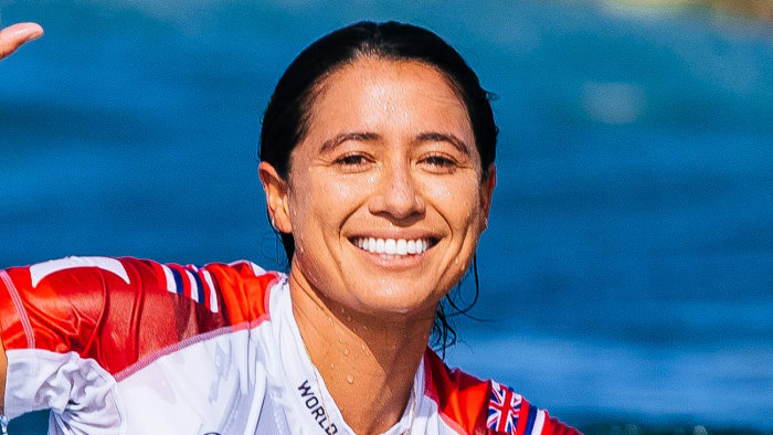 Malia Manuel sits atop a surfboard in the water and smiles for the camera.